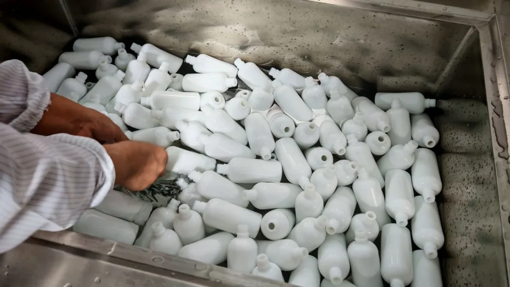 Hands manually cleaning white glass cosmetic bottles in a sterile sink, ensuring strict hygiene for professional OEM filling.