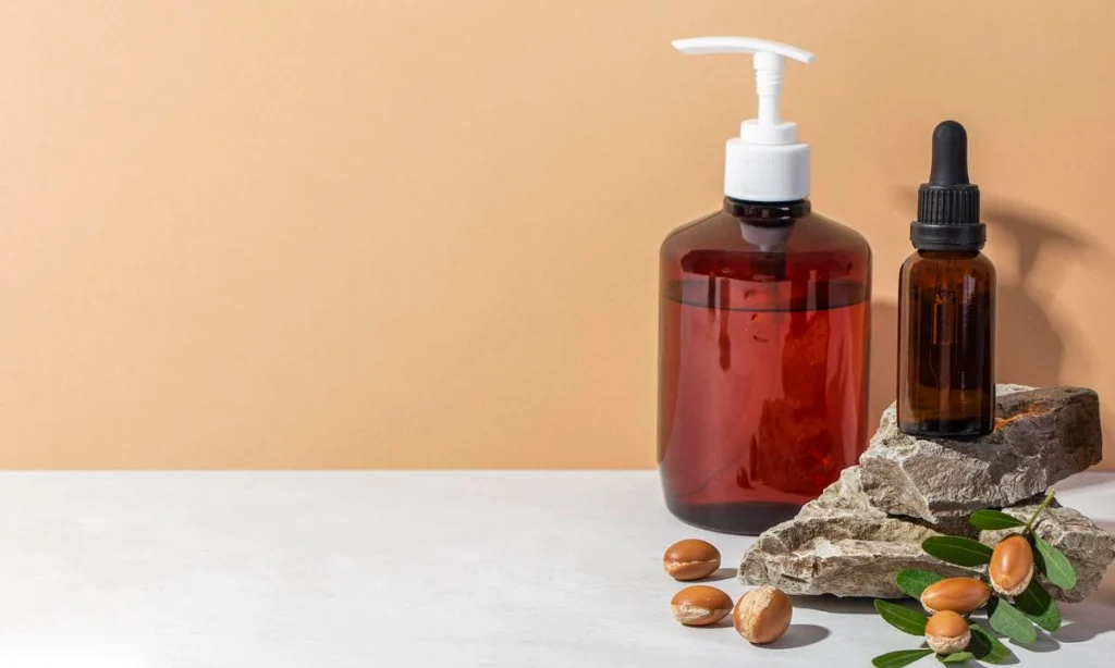 An argan oil lotion pump bottle and a dropper bottle on natural stone, with fresh leaves.