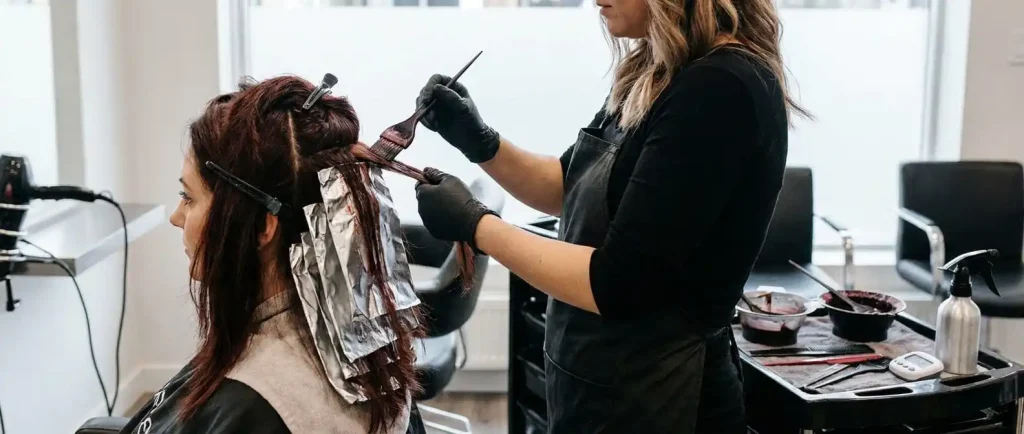 Close-up of a stylist applying dark hair dye and using foil highlights on a client in a salon setting.