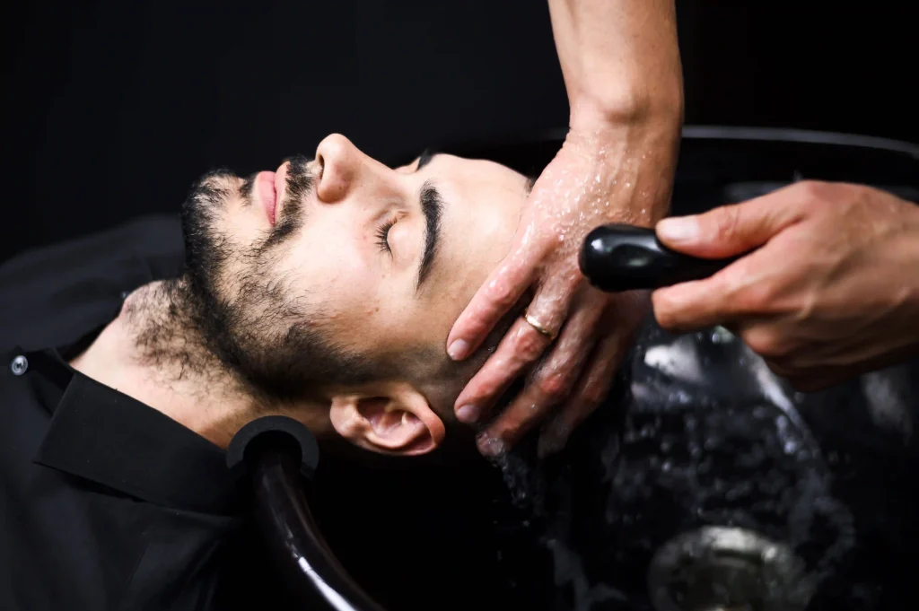 Close-up of a stylist washing a bearded man's hair in a professional black salon basin.