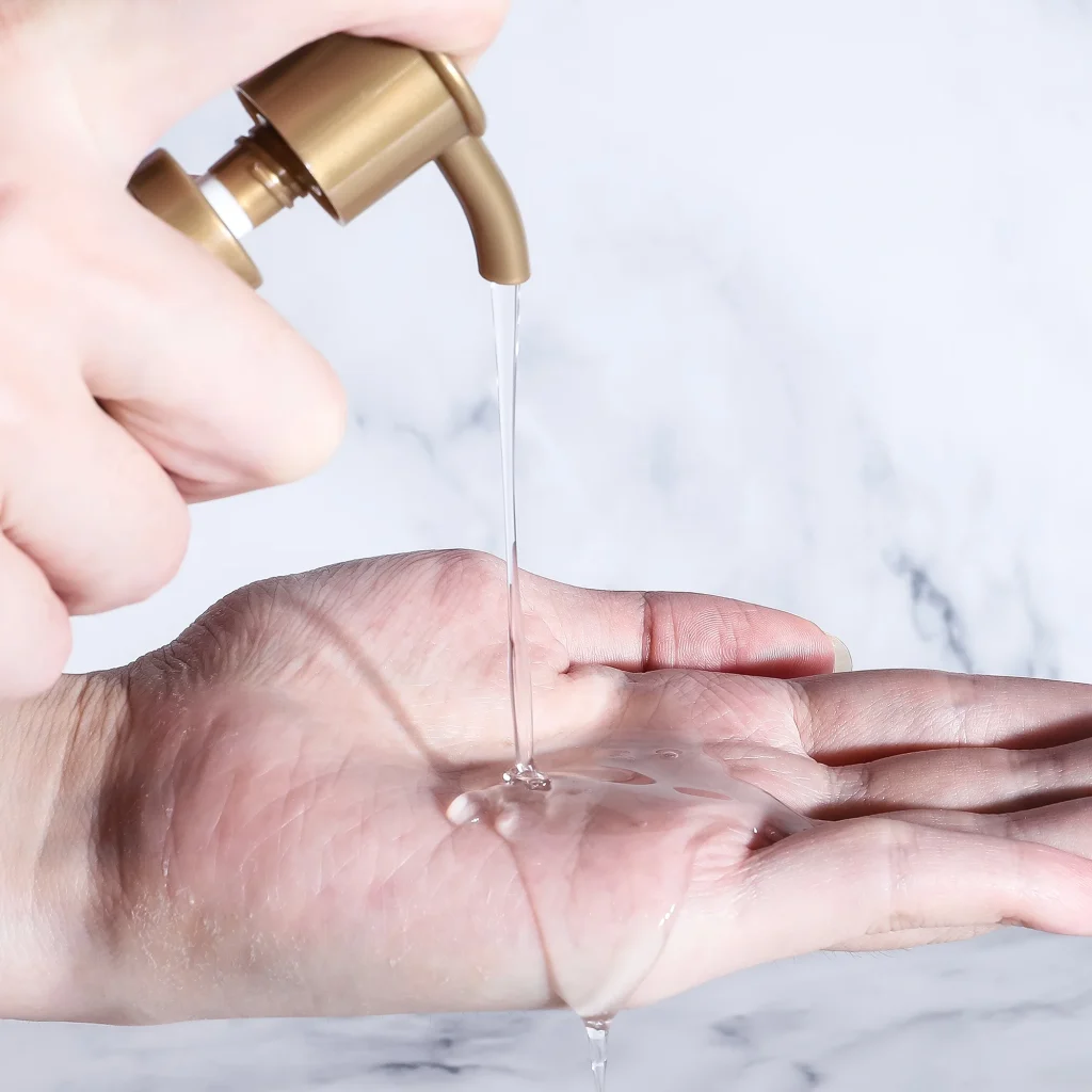 A close-up shot of a hand receiving clear, liquid clarifying shampoo dispensed from a gold pump, set against a white marble background.