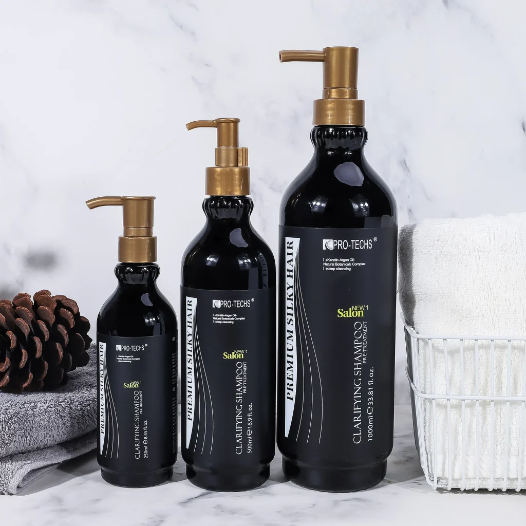 Three black bottles of "CLARIFYING SHAMPOO" in 250ml, 500ml, and 1000ml sizes with gold pump dispensers, arranged by height on a marble surface next to a pinecone and towels.