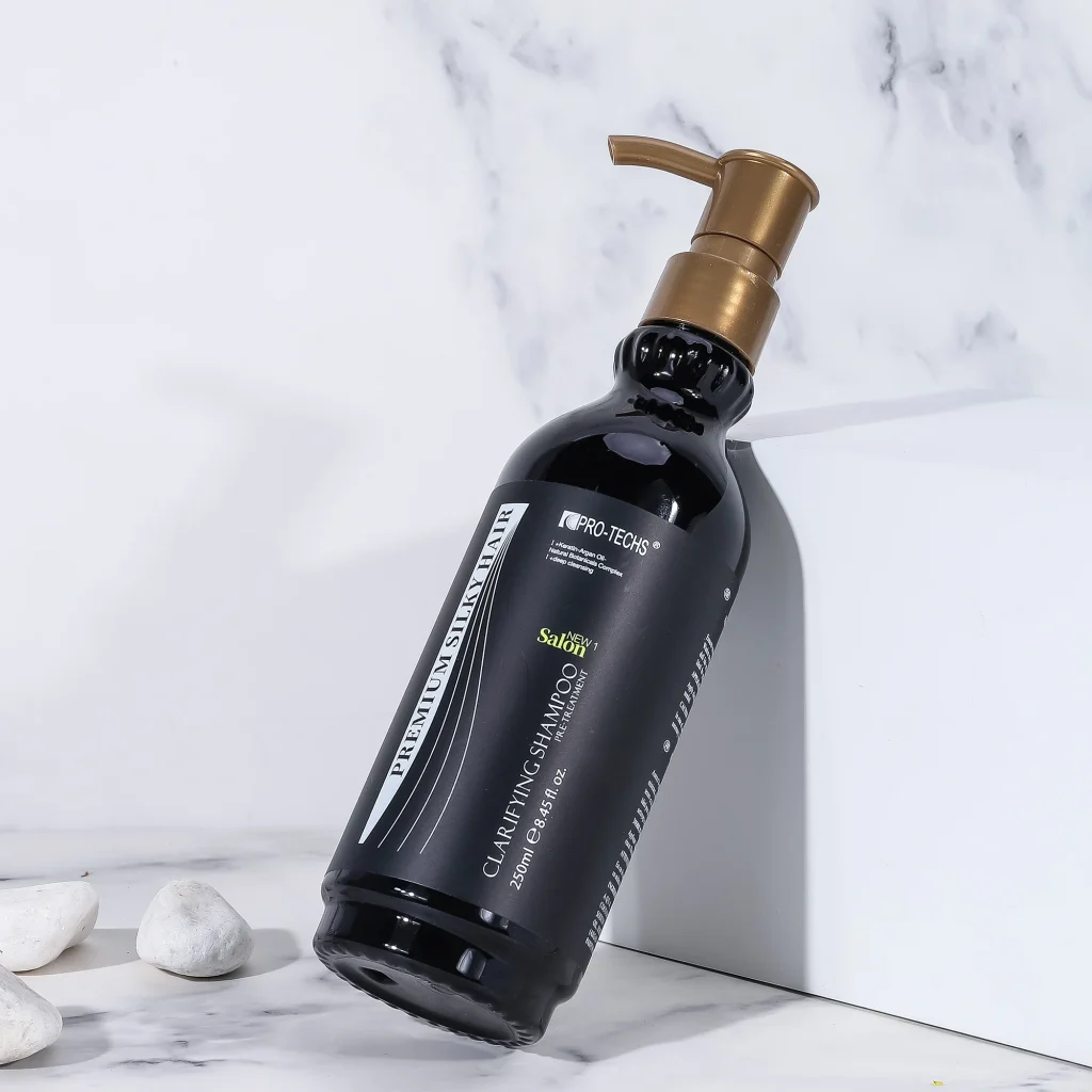 A sleek black 250ml bottle of "CLARIFYING SHAMPOO" with a gold pump dispenser, leaning against a white block on a marble surface with decorative white stones.