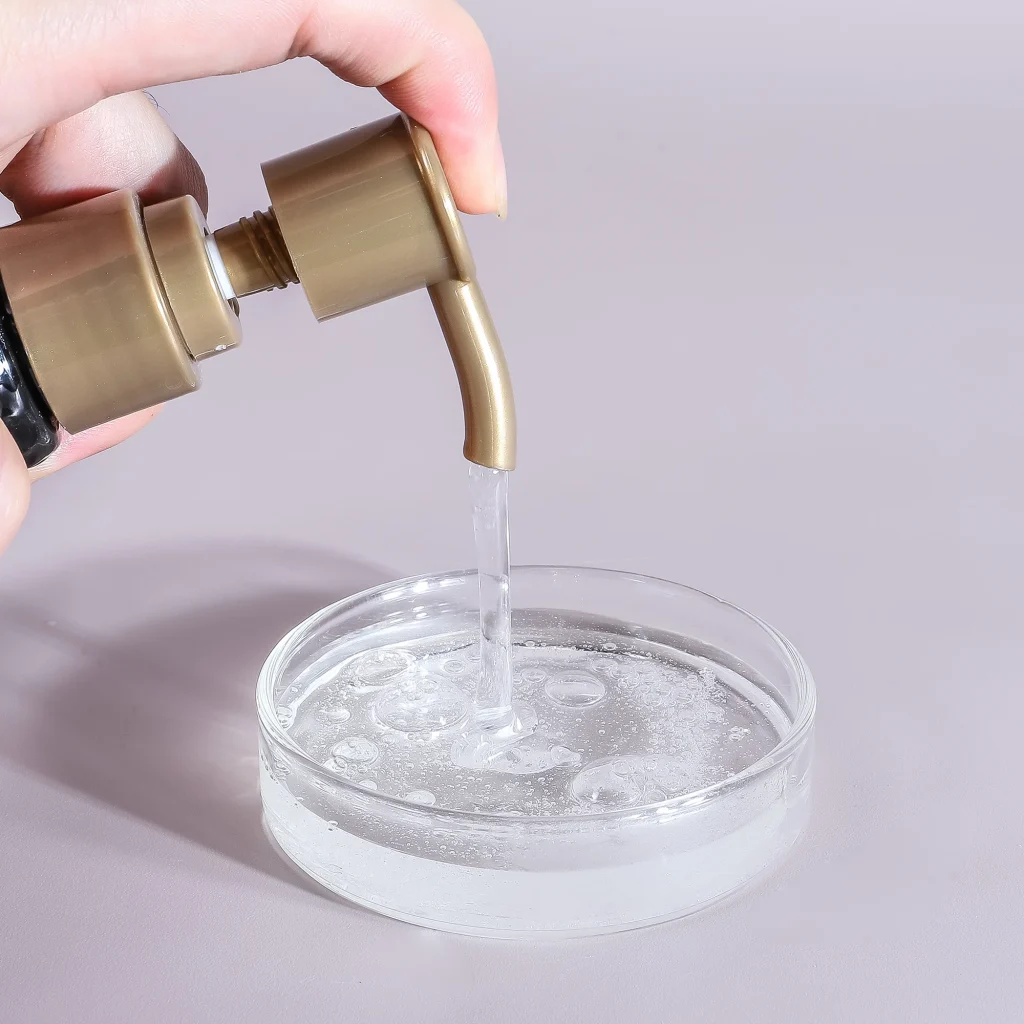 A person's hand pressing a gold pump to dispense clear, viscous clarifying shampoo into a small glass dish, highlighting its transparent and fluid texture.
