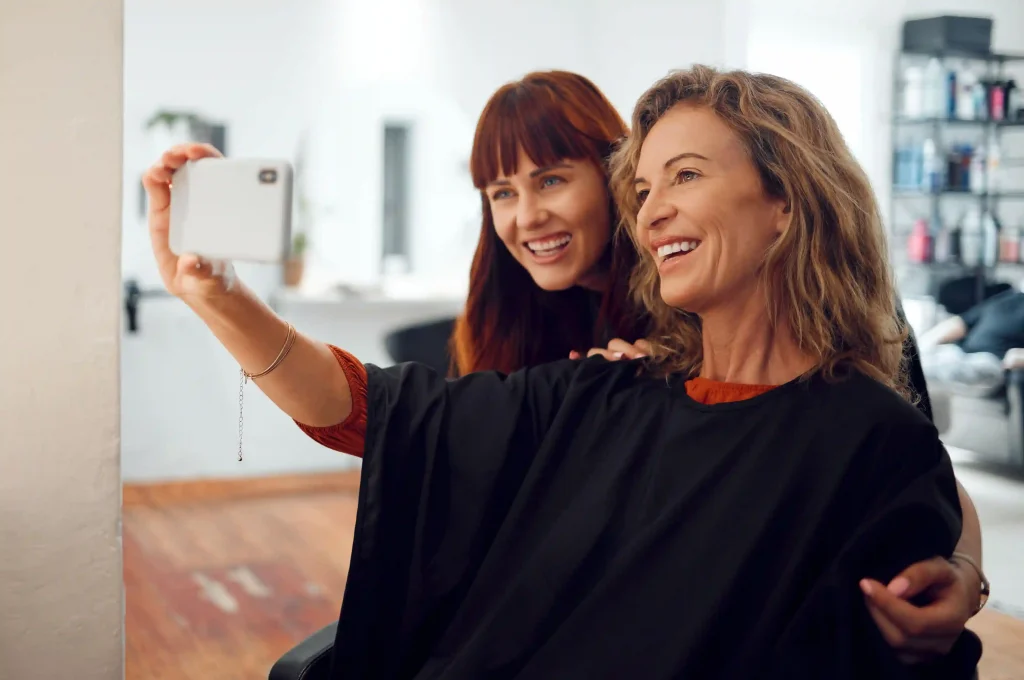 Happy client taking a selfie with her stylist in the mirror to show off her new voluminous haircut.