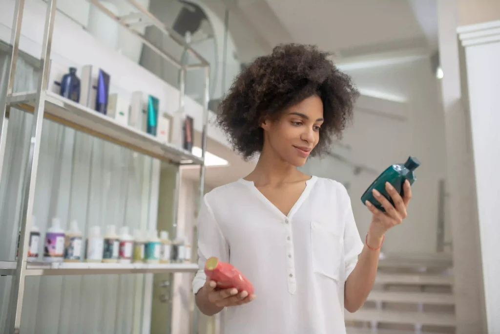 A woman in a store comparing two different hair care bottles while looking at a stocked shelf.