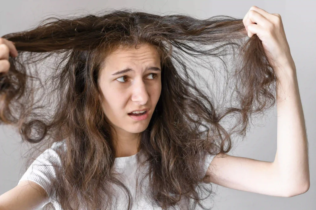 A woman looking frustrated while holding her dry, frizzy, and tangled brown hair.