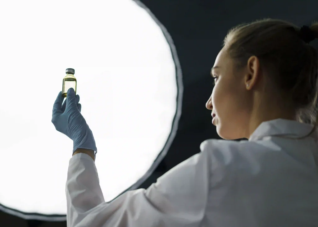 A scientist in a white coat and gloves inspecting a small vial of hair oil against a bright light in a lab setting.