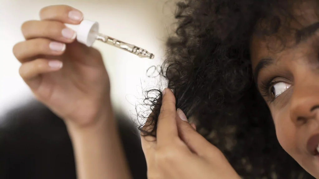 Close-up of a person applying hair oil with a dropper to curly hair, demonstrating product texture and use.