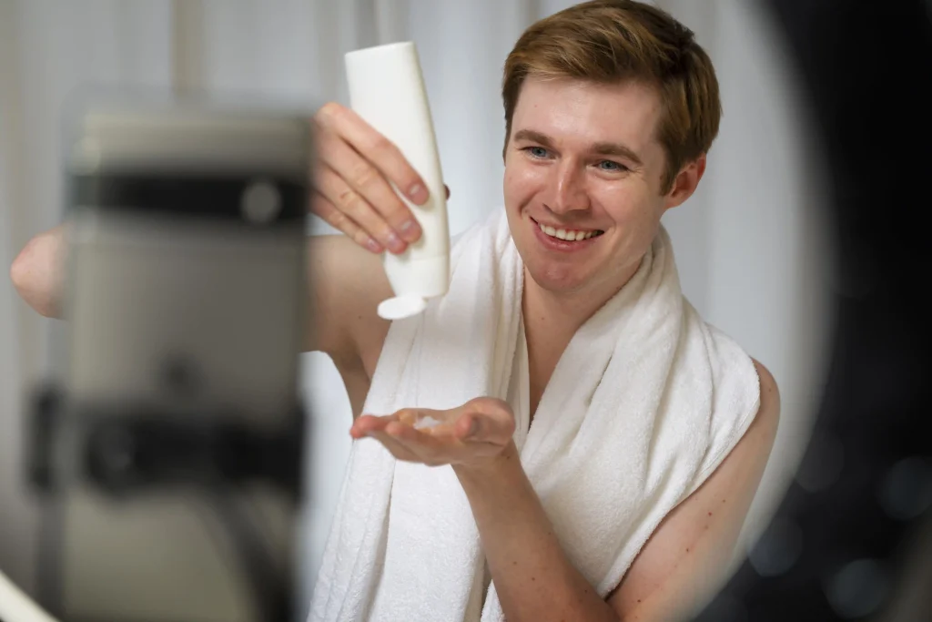 A smiling man in a towel applies hair cream while filming a tutorial on his smartphone with a ring light.