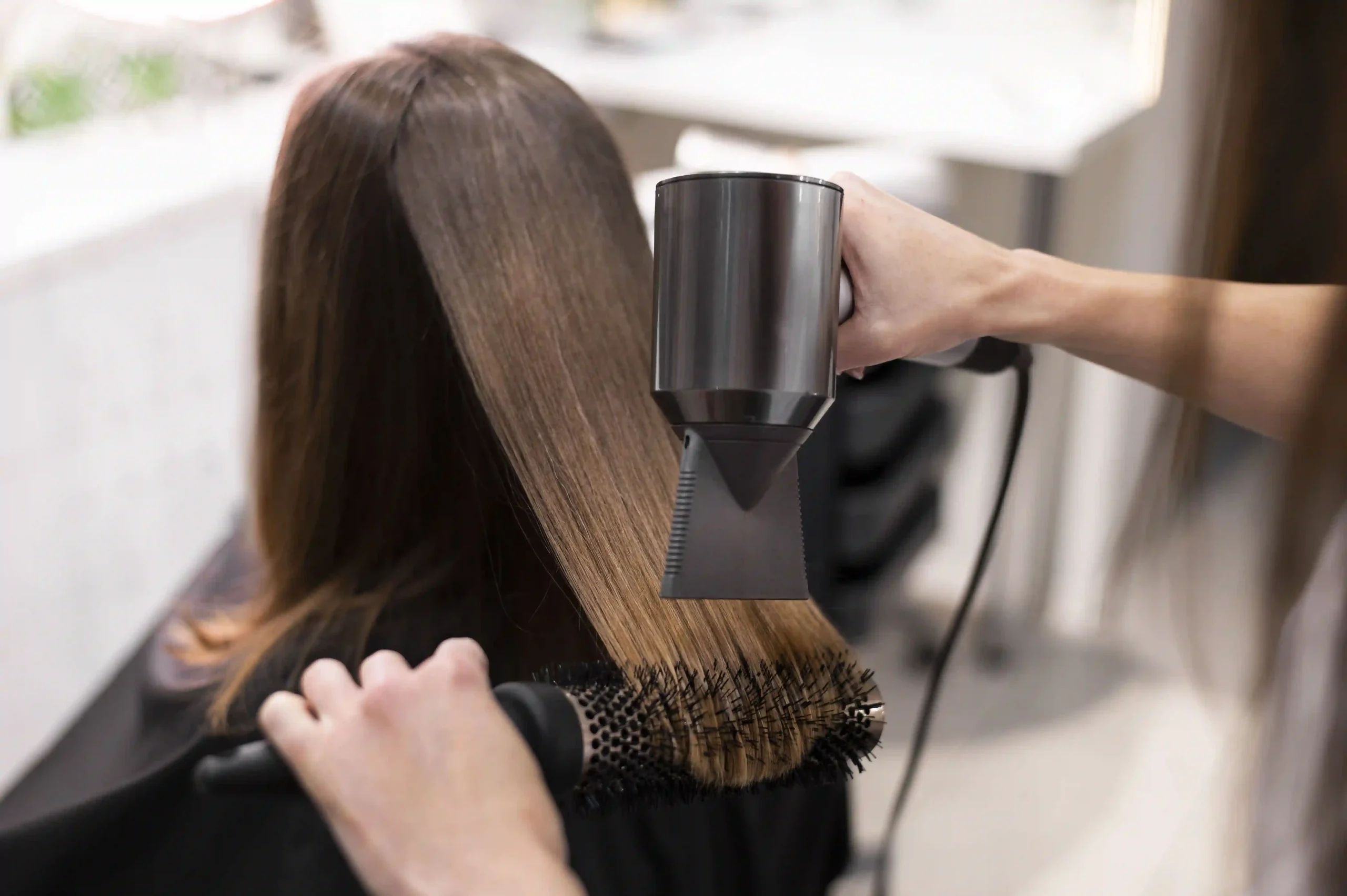 Professional stylist blow-drying a woman's long, smooth brown hair into a sleek finish in a bright salon.