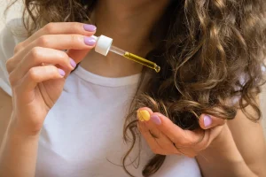 Woman with fine curly hair applying a drop of yellow hair oil from a glass dropper to her ends.