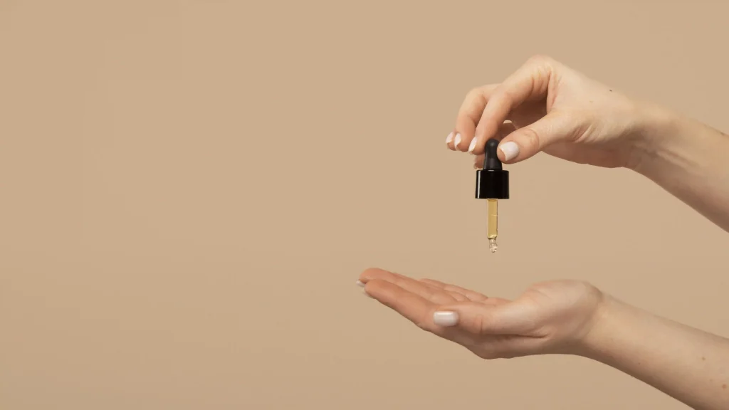 Close-up of hands holding a glass dropper over an open palm, preparing to apply a golden hair serum.