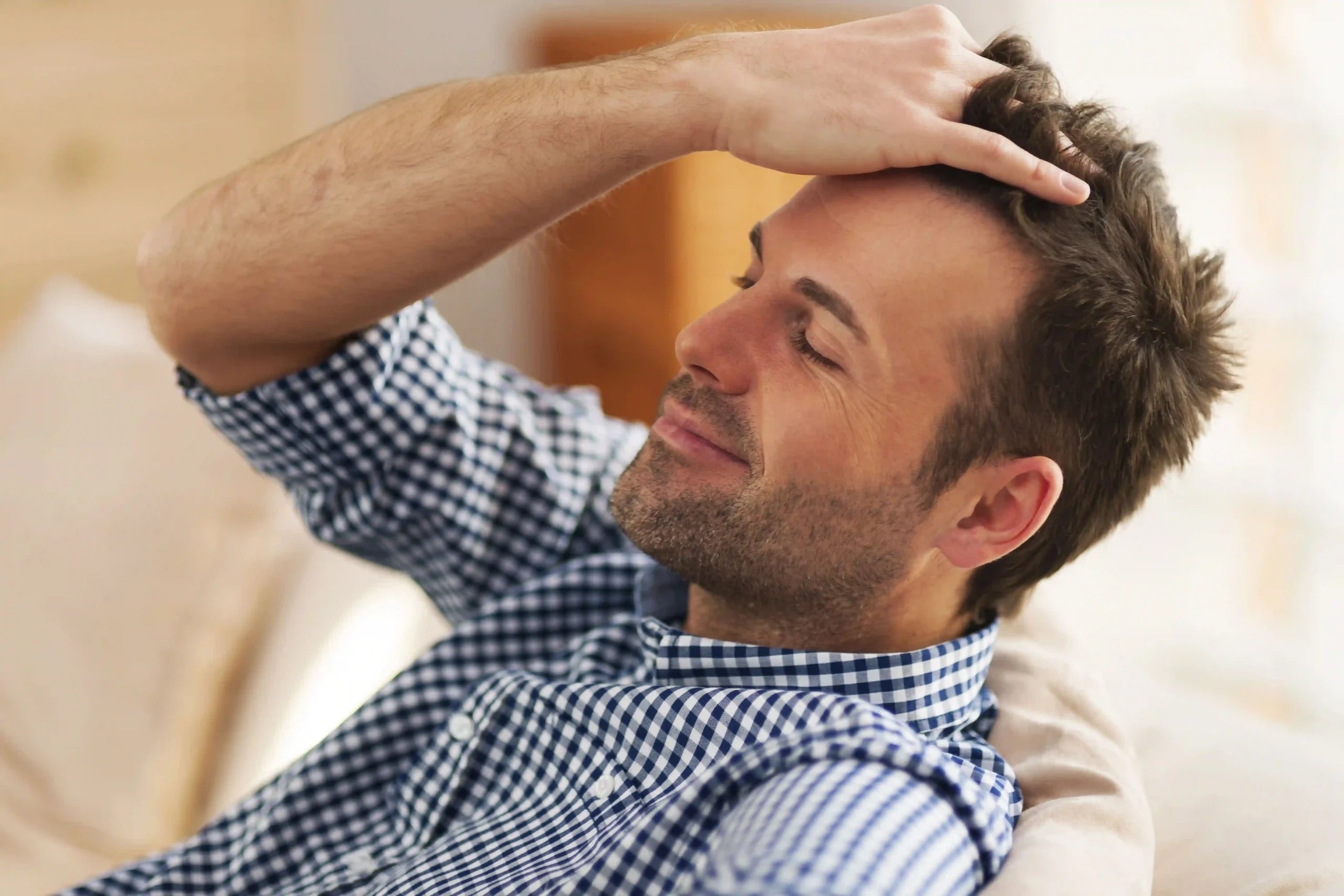A relaxed man in a checkered shirt smiles with eyes closed while touching his well-groomed hair.