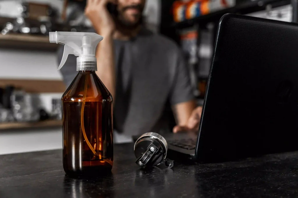 A spray bottle in focus on a desk while a man works on a laptop in the blurred background.