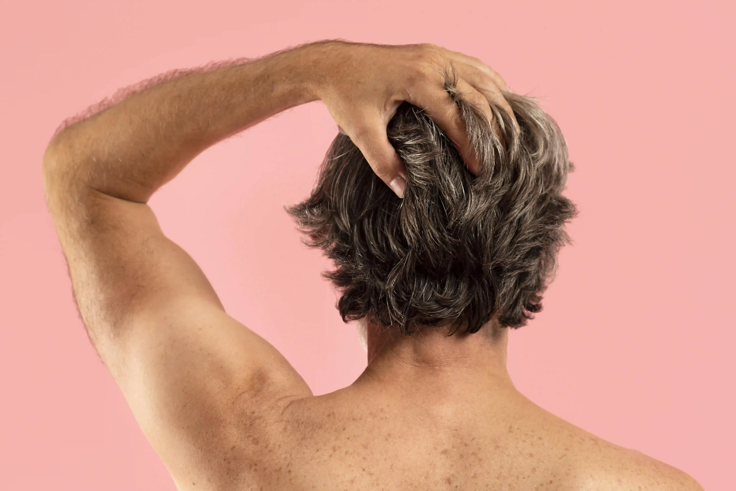 Rear view of a man with thick, textured salt-and-pepper hair against a soft pink background.