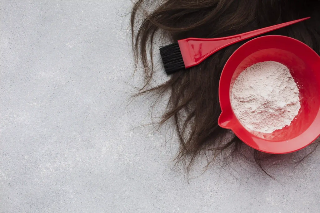 A red mixing bowl with white powder and a hair dye brush next to dark hair strands.