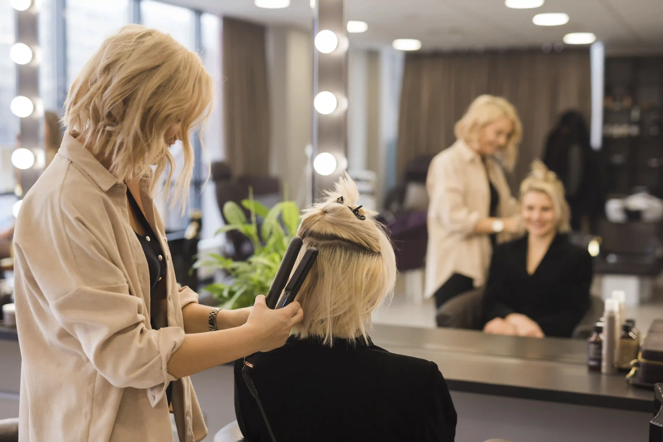 Stylist using a flat iron on a blonde client's hair in a modern, bright hair salon setting.