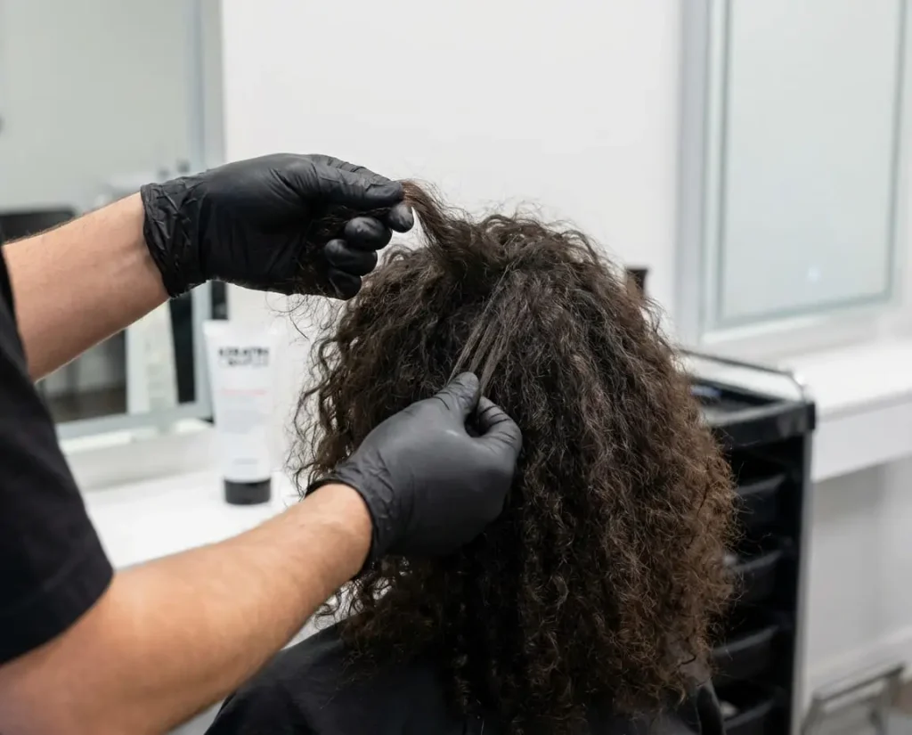 A stylist in black gloves examines a client's natural curly, frizzy hair before a keratin treatment.