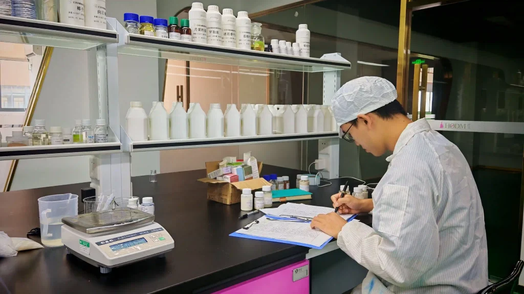 A scientist in a lab coat and hairnet recording finished product inspection data on a clipboard at a laboratory desk.