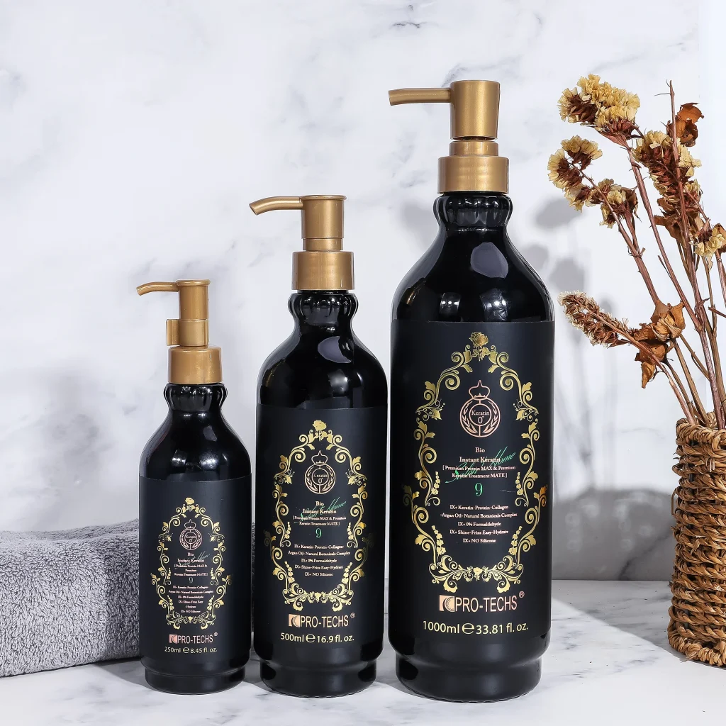 Three black bottles of "Keratin Bio Instant 9" hair treatment in 250ml, 500ml, and 1000ml sizes with ornate gold labels and pump dispensers, arranged by height on a marble surface next to a grey towel and dried flowers.