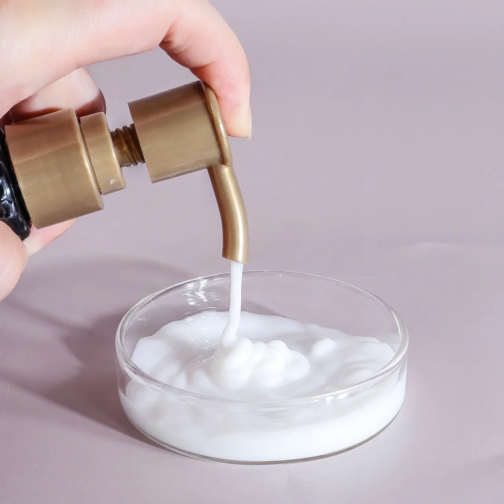 A close-up shot of a hand dispensing thick, white "Renewal Keratin Smooth Conditioner 4" from a gold pump into a glass dish, highlighting its rich and creamy texture.