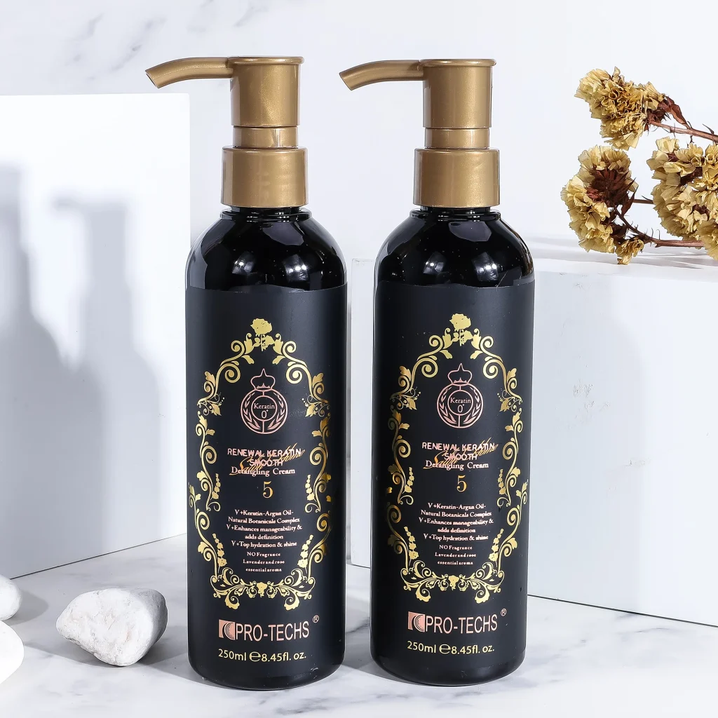 Two 250ml black bottles of "Renewal Keratin Smooth Detangling Cream 5" with gold pumps and ornate gold labels, standing side-by-side on a marble surface next to white stones and dried yellow flowers.