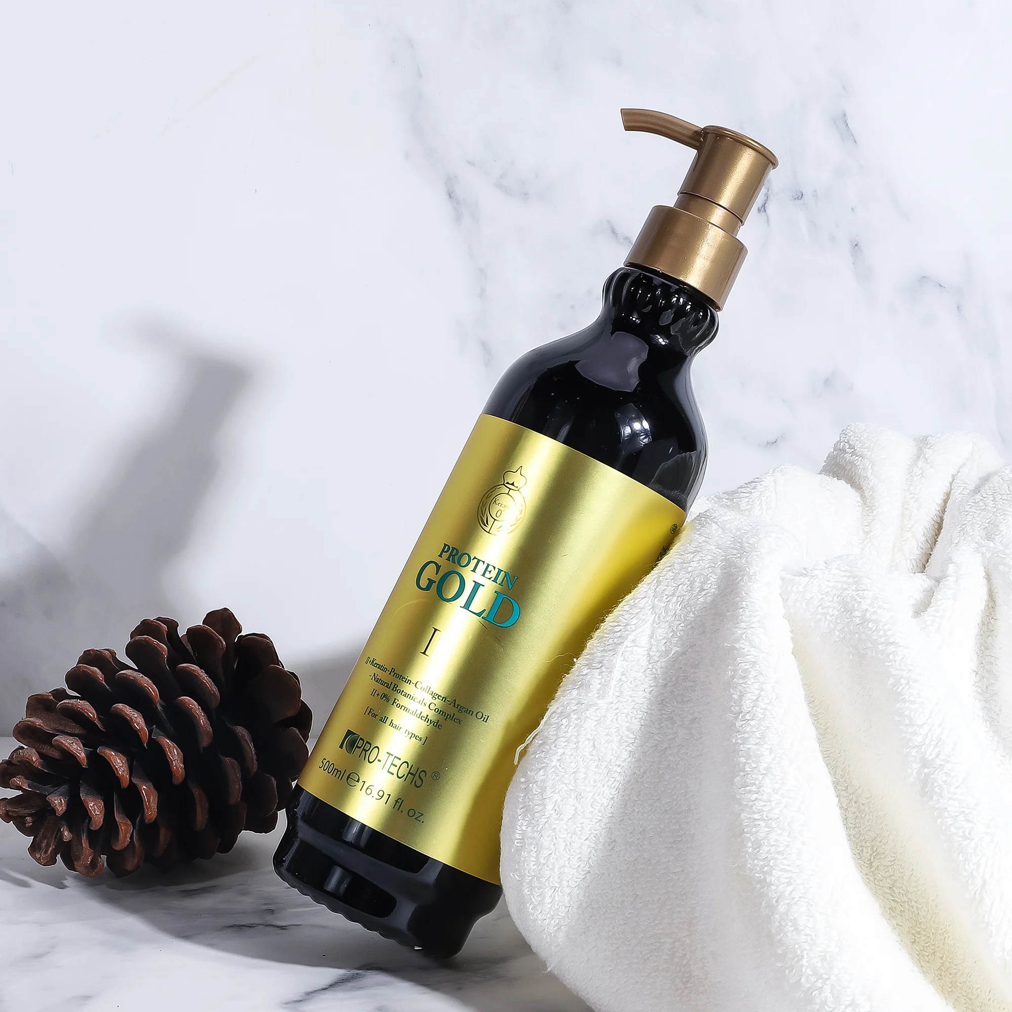 A black 500ml bottle of "PROTEIN GOLD II" hair treatment with a metallic gold label and pump, resting against a white towel next to a pinecone on a marble surface.