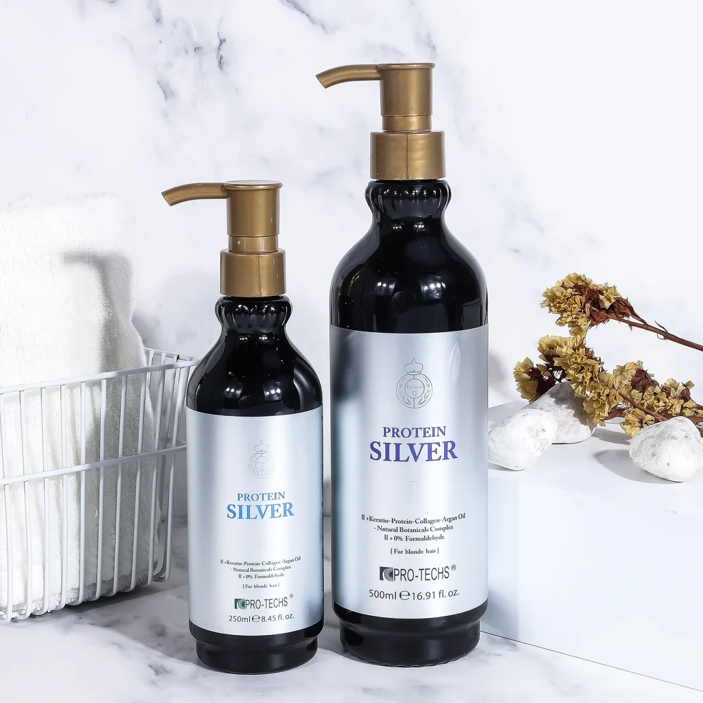 Two black bottles of "Protein Silver II" hair treatment (250ml and 500ml) with silver labels and gold pumps, displayed on marble with a pinecone, towel, and stones.