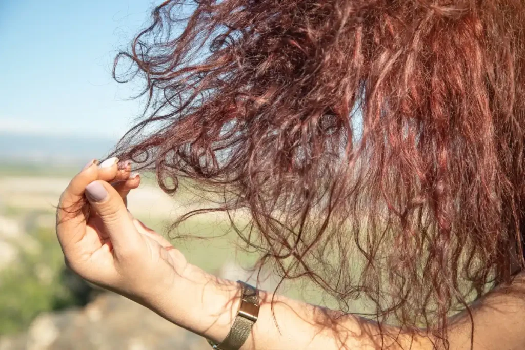 Close-up of a person's hand touching dry, reddish-brown curly hair with visible frizz and flyaways against a bright outdoor background.