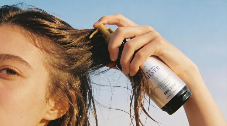 A close-up of a woman applying Pro-Techs Protein Silver hair product to her damp brown hair against a clear blue sky background.