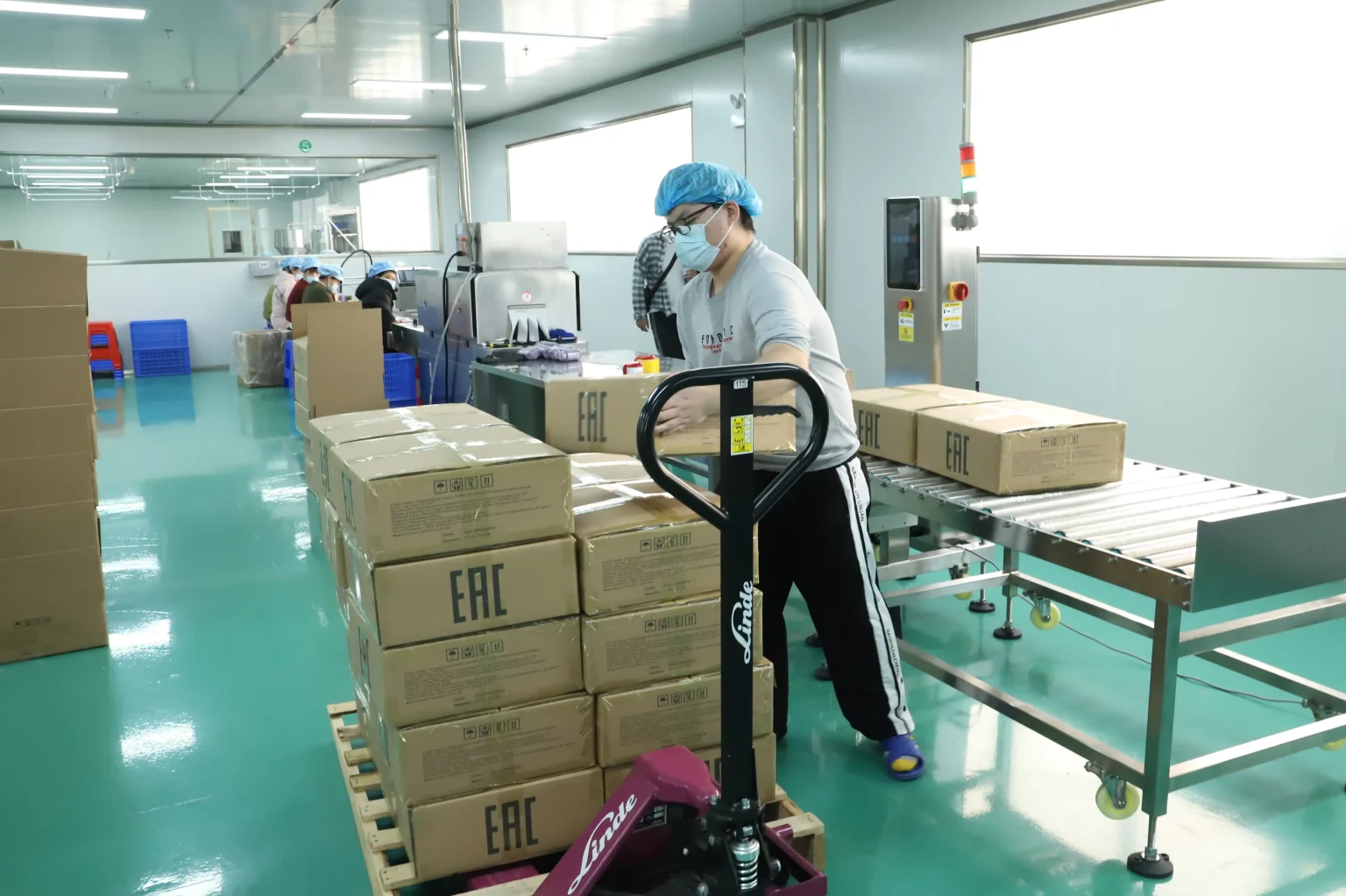 Factory workers wearing blue caps and masks assemble boxes on a packaging line with stacked cartons nearby.
