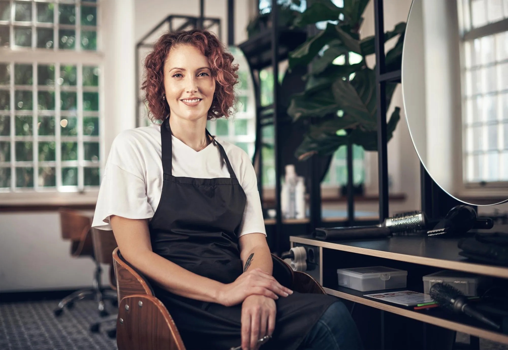 Smiling woman with curly red hair in a white shirt and black apron sits in a salon chair by a mirror.
