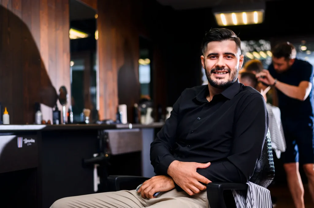 Smiling man in a black shirt seated in a barber chair, while a barber works on another client in the background.