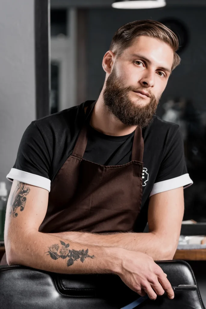 Bearded man in a brown barber apron leaning on a chair with a straight razor in hand inside a barbershop.