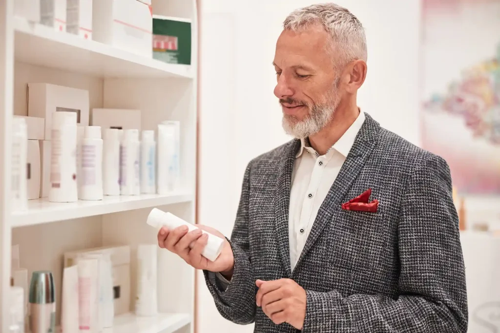 Older man with gray beard in a checked blazer examines skincare products on a shelf, holding a white tube.