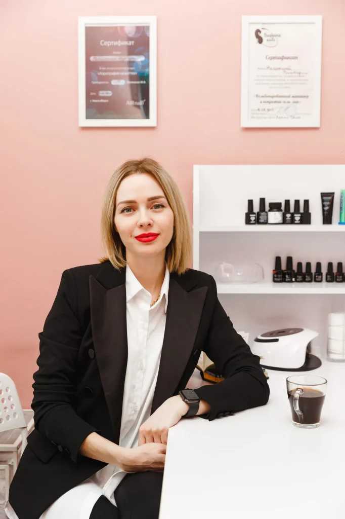 Professional woman in a black blazer sits at a white desk in a pink-walled salon, with nail products on a shelf behind her.