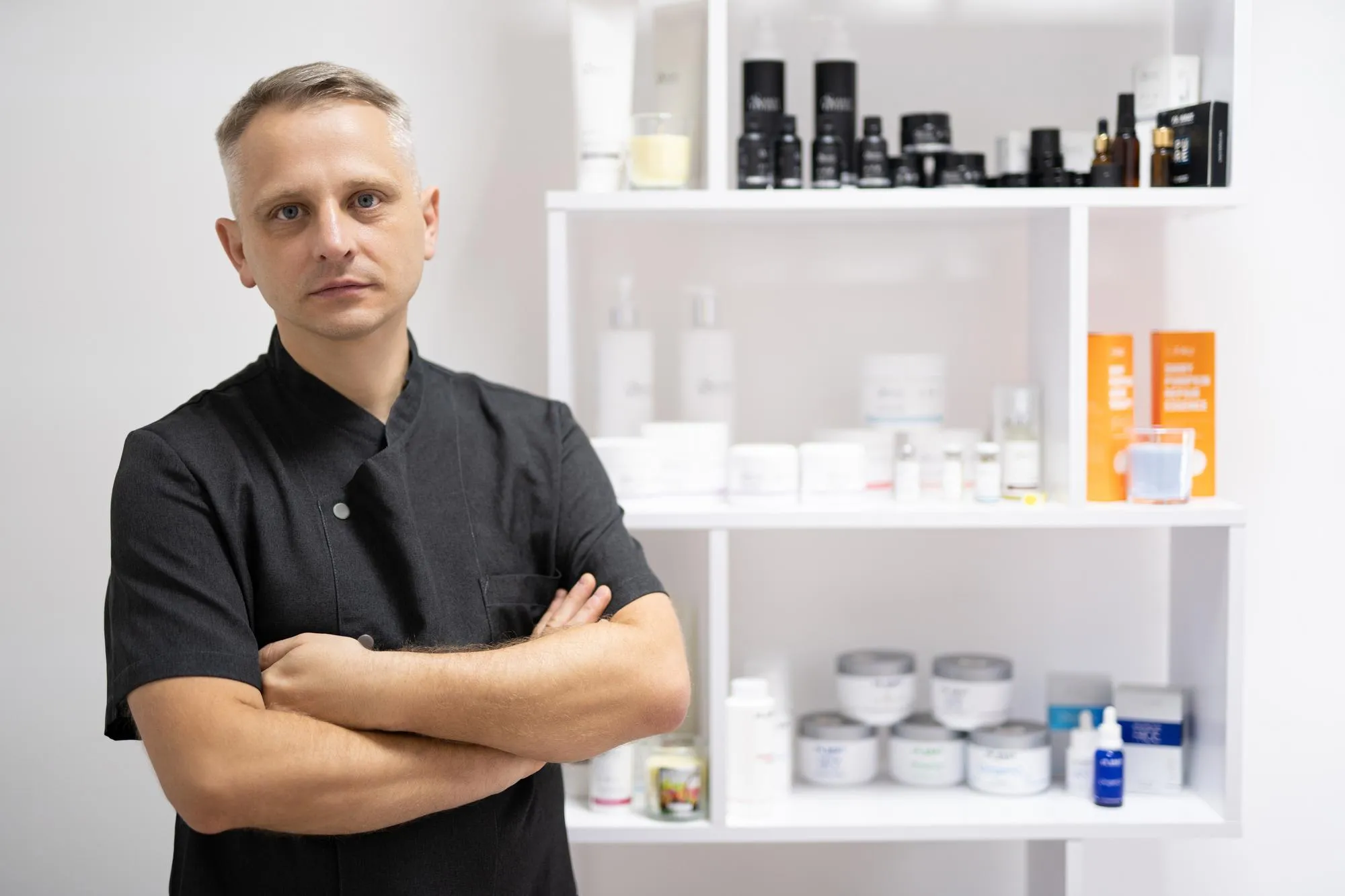 Man in a black uniform stands with arms crossed in front of white shelving filled with skincare bottles and boxes.