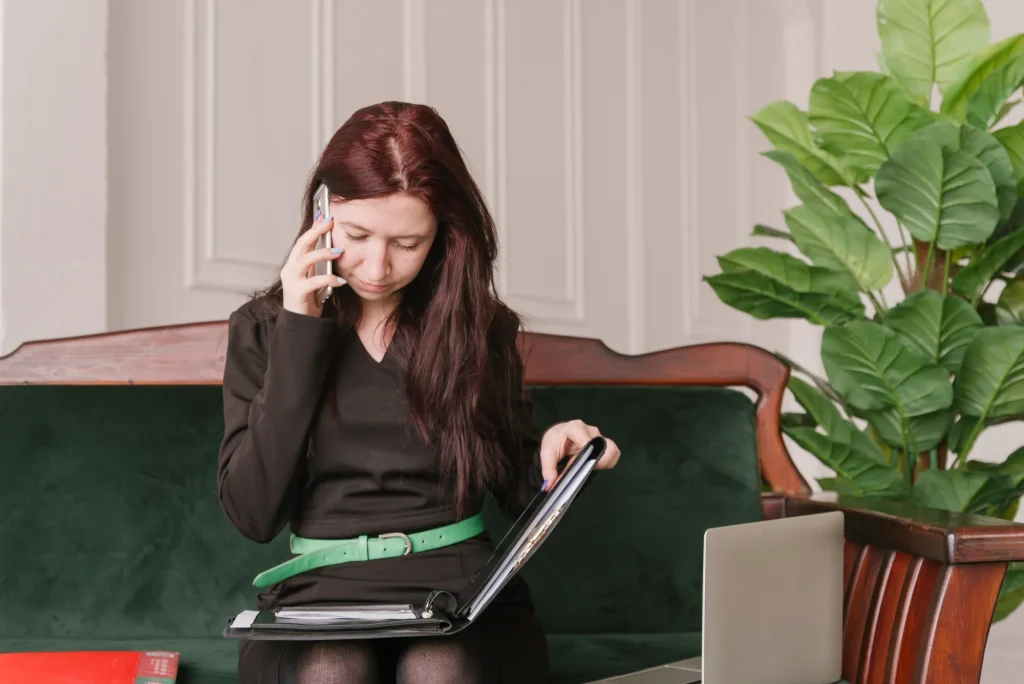 Woman with dark hair in a black outfit sits on a green velvet sofa, talking on a mobile phone while holding a folder; a large leafy plant is on the right.