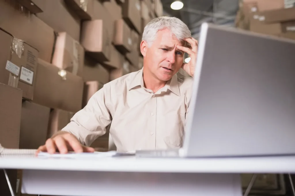 Middle-aged man in a warehouse, hand on head, focusing on a computer screen with tall cardboard boxes in the background (stress or confusion)