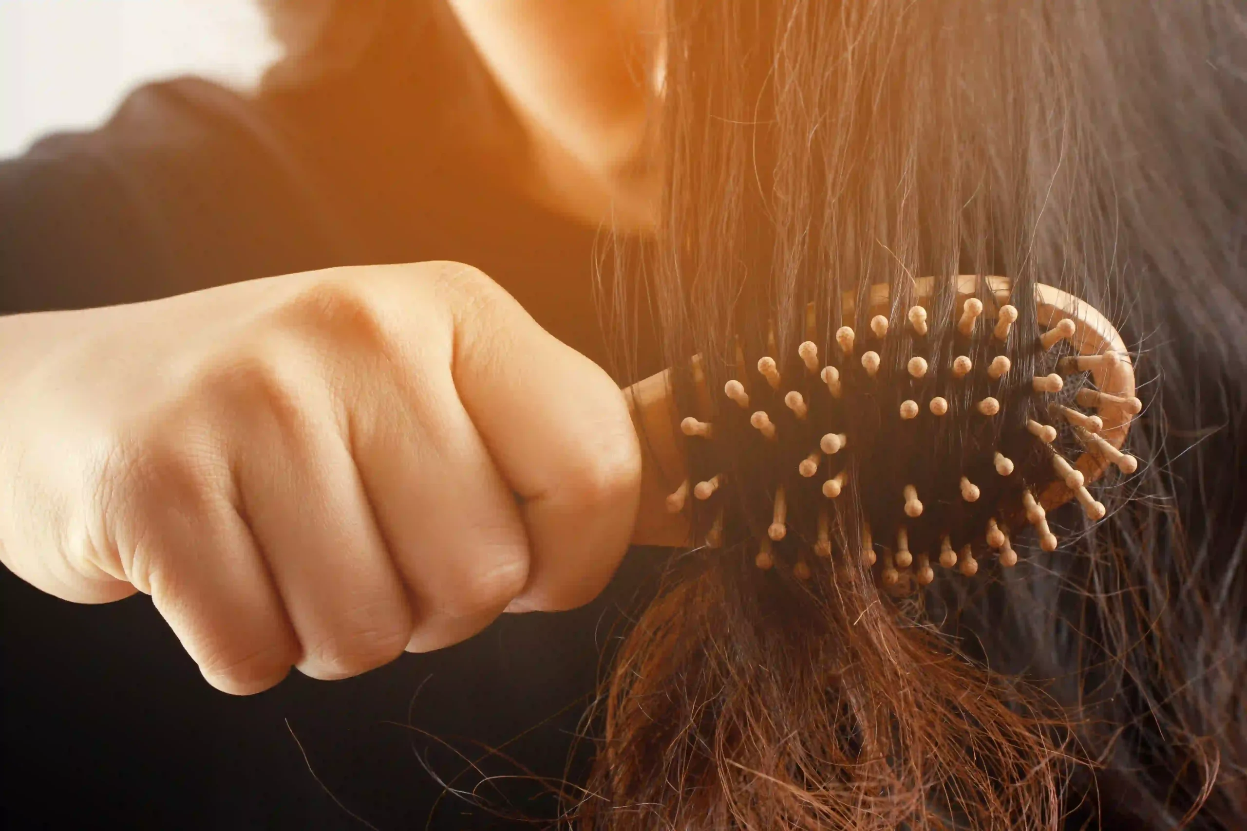 Rear view of a woman brushing tangled hair with a wooden comb, illustrating hair loss and breakage concerns.