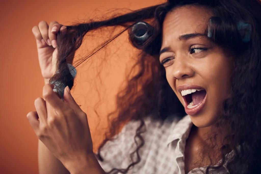 A young woman with a pained expression struggling to remove blue rollers from her tangled frizz hair against an orange background.