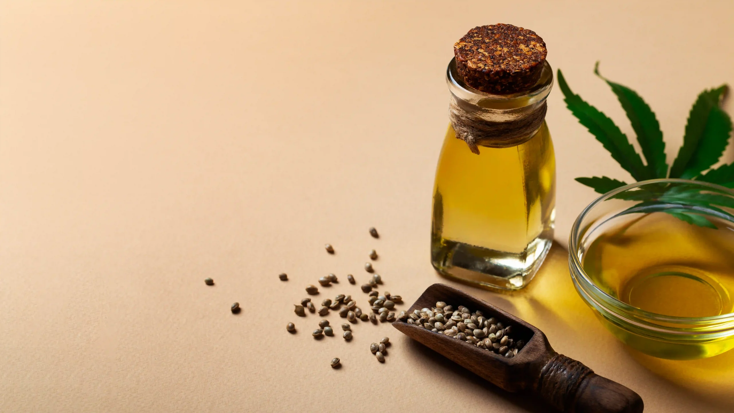 A corked glass bottle of golden castor oil and a glass bowl of oil, shown with hemp seeds on a wooden scoop and a green leaf on a tan background.