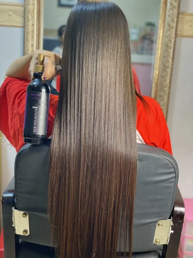 Back view of a person in a salon chair with long, straight, shiny brown hair, holding a dark bottle of keratin treatment.