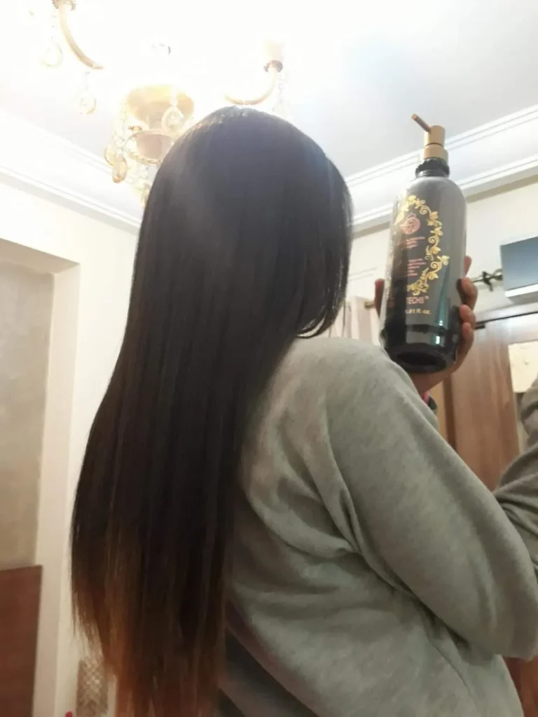 Back view of a person with long dark hair wearing a gray sweatshirt, holding a tall dark bottle with a pump dispenser indoors.