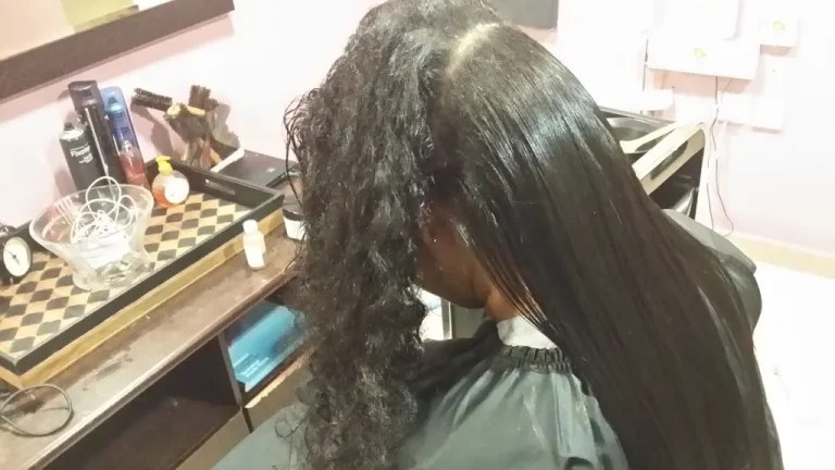 Person seated in a salon chair with one side curly and the other straightened during a hair treatment in a beauty shop.