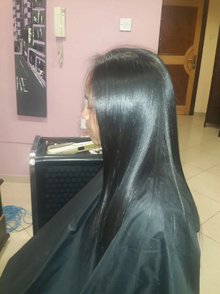 Side profile of a person with long, straight black hair wearing a black salon cape in a hair salon.