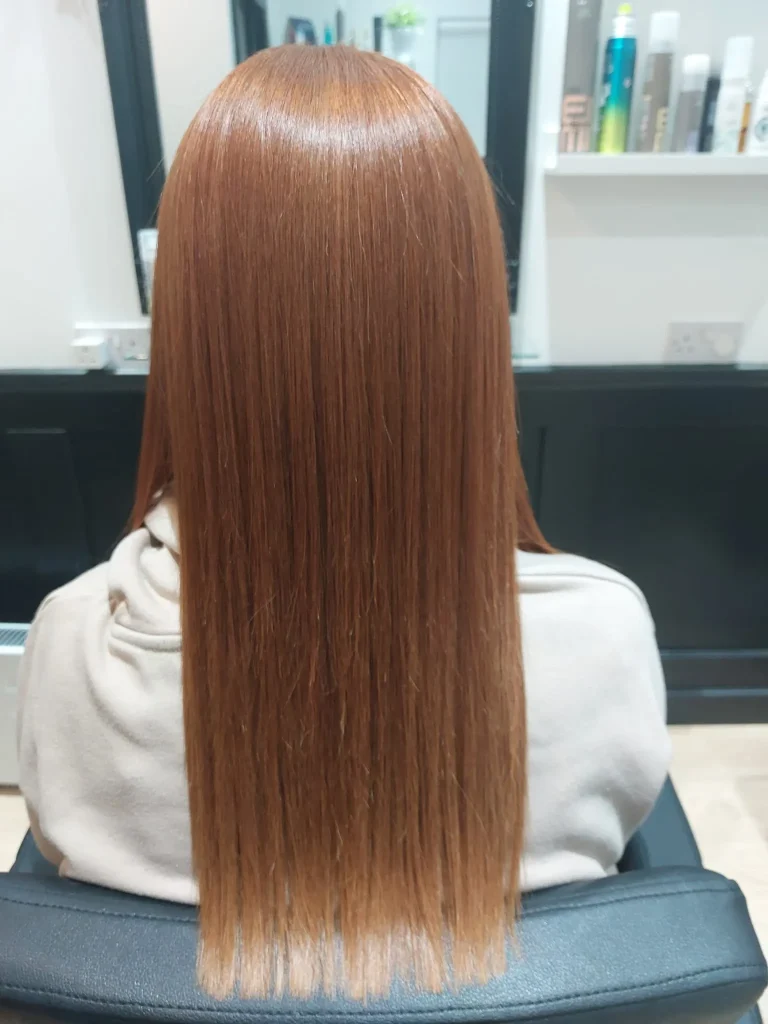 Back view of a person with long straight copper-red hair in a salon chair, shelves and mirror visible in the background.