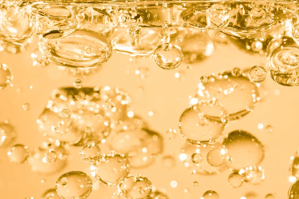 An abstract close-up of numerous golden oil bubbles of varying sizes suspended in a clear liquid, creating a shimmering, textured effect.