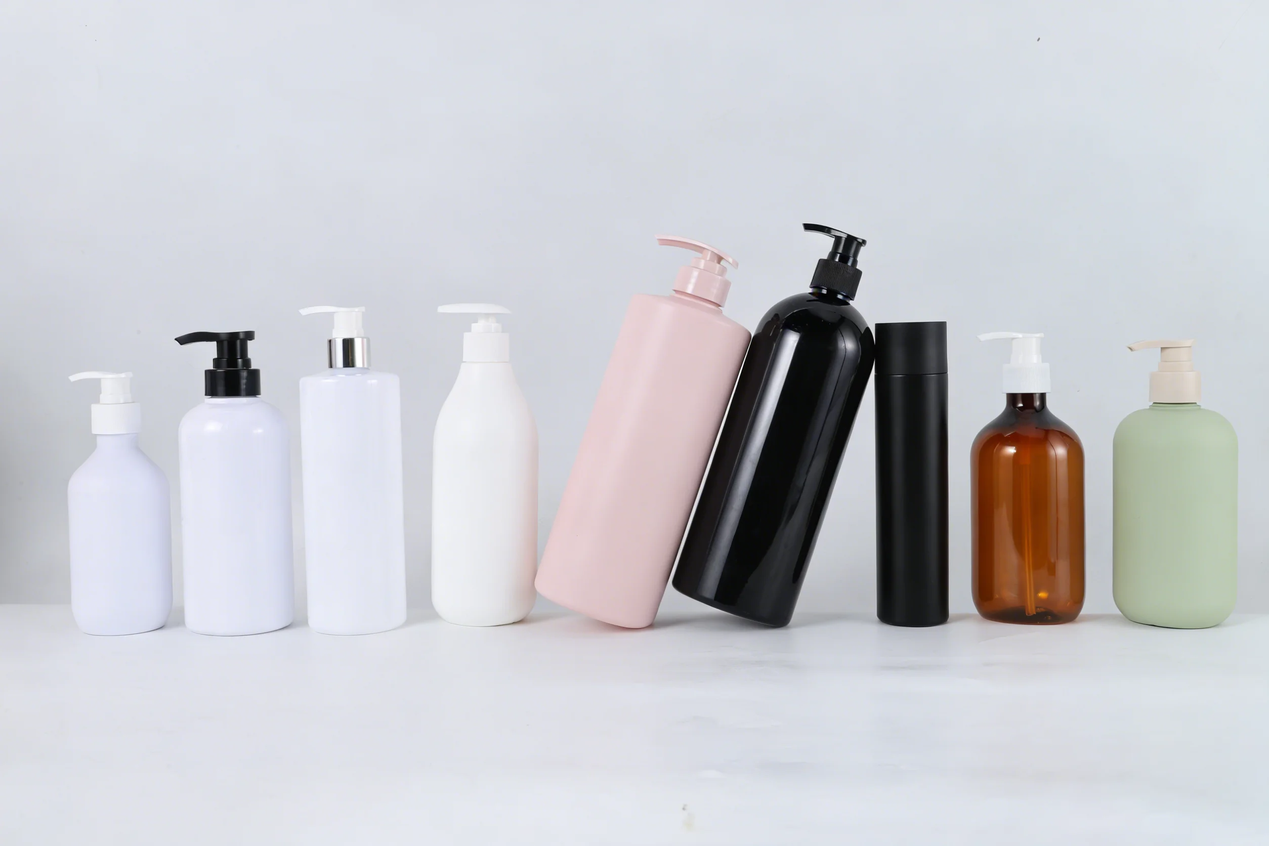 A row of nine assorted blank hair care pump bottles and containers in white, pink, black, amber, and light green colors on a white surface.